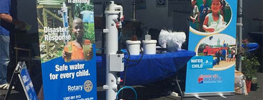 A table with a blue cloth holds water sanitation equipment. A large water sanitation pipe machine stands in front of the table. Rotary’s safe water pull-up banners sit on either side of the table.