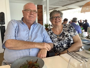 Jenny and Trevor  are sitting smiling in the Kingfisher Grove community centre. Trevor wears a blue and white checked shirt, and Jenny wears a black and white floral printed top.