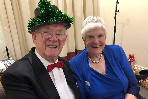 John and Wilma at Burwood Terrace. John wears a black suit, a red bowtie and a celebratory hat. Wilma wears a blue dress, stylish jewellery and a sparkly tiara.