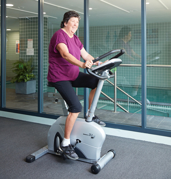 Jill is working out on an exercise bicycle and through the glass wall behind her is the community pool.
